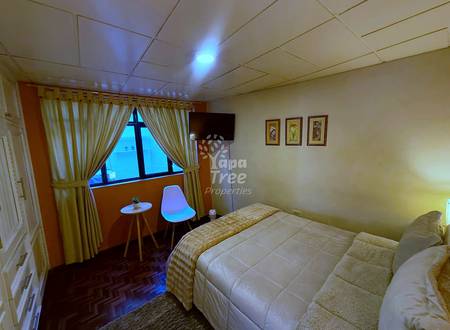 Bedroom with two single beds, wall-mounted TV, window with cream curtains, small wooden table, white chair, and framed botanical prints on beige walls.
