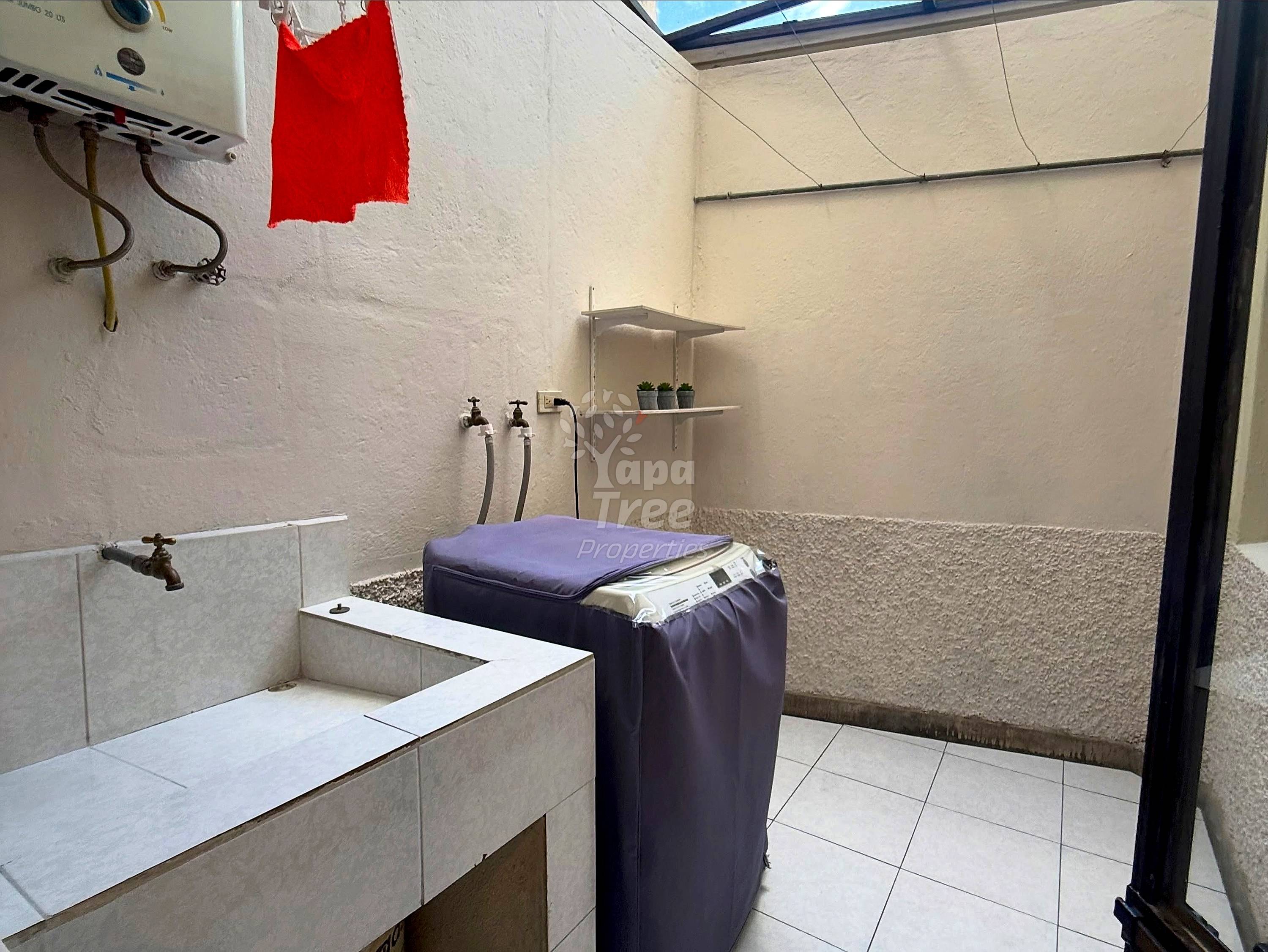 Bathroom with white tile floor, concrete sink basin, wall-mounted faucet, shelving, and washing machine under skylight.