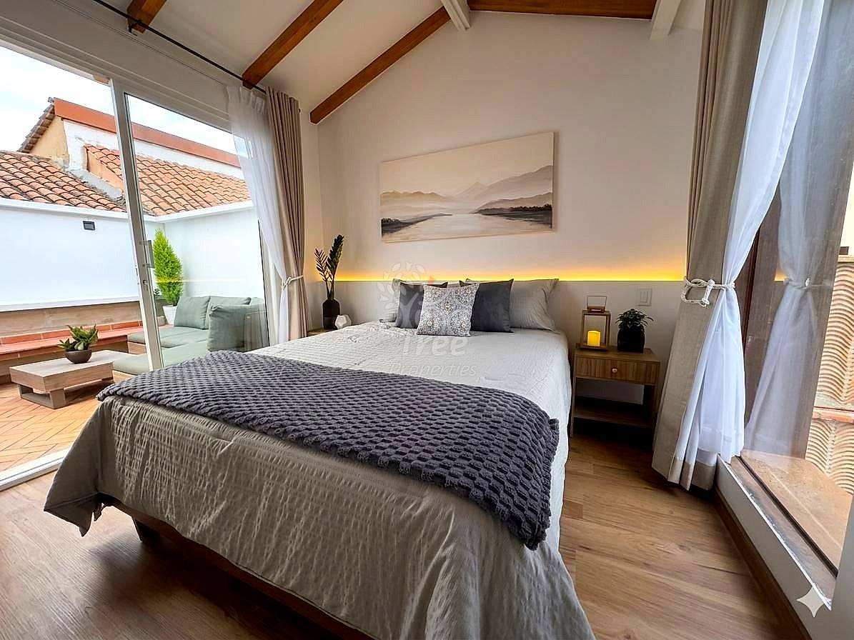 Bright bedroom with queen bed, wooden beams, warm accent lighting, wood nightstand, glass doors opening to terrace with tile roof views.