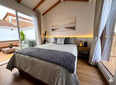 Bright bedroom with queen bed, wooden beams, warm accent lighting, wood nightstand, glass doors opening to terrace with tile roof views.