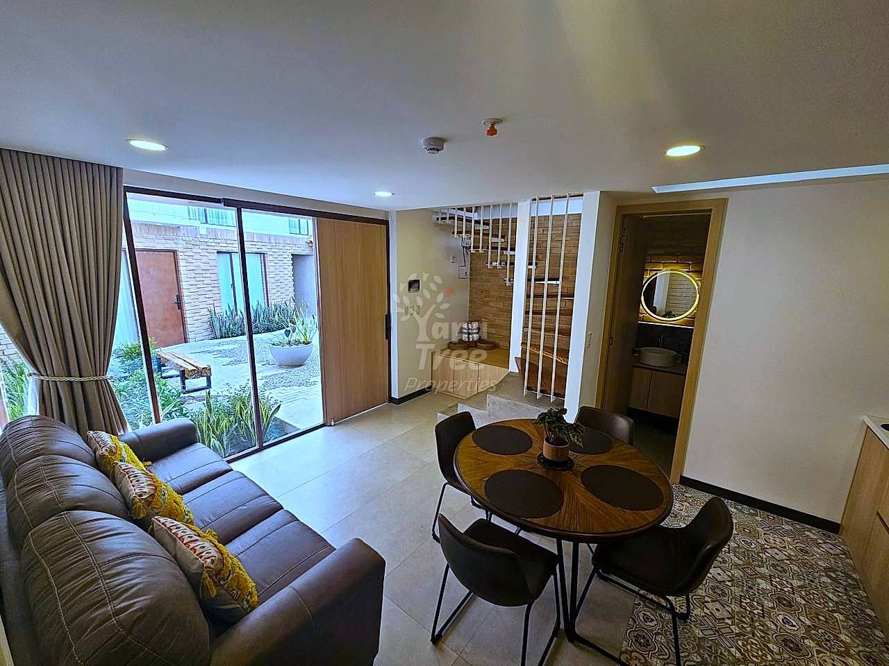 Living and dining area with brown leather sofa, round wooden table with black chairs, tile flooring, glass patio doors, staircase, and recessed ceiling lights.