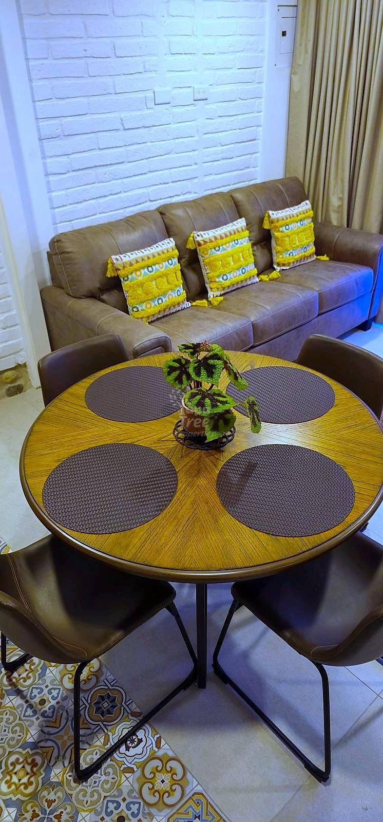 Living room with brown leather sofa, yellow patterned pillows, round wood dining table with four brown chairs, and white brick wall.