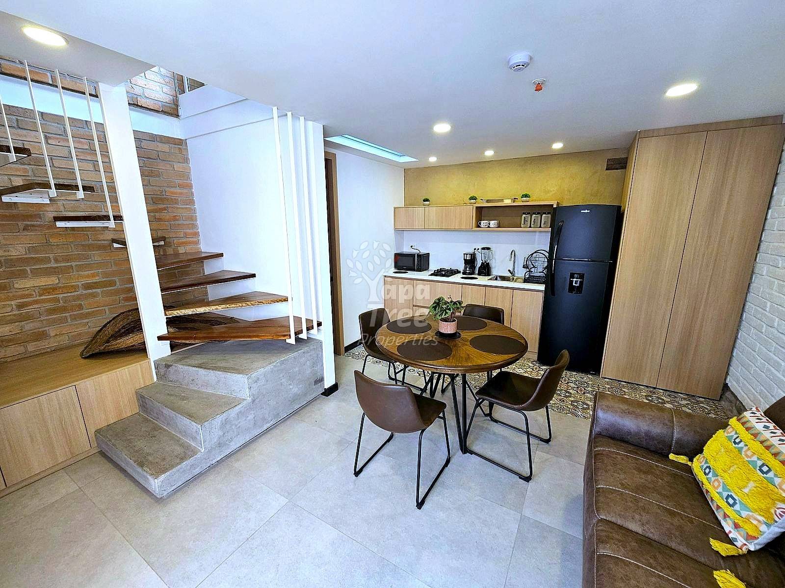 Open-plan departamento with dining table, wood and brick staircase, integrated cocina with black refrigerator, and recessed ceiling lights