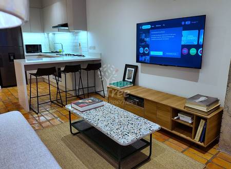 Open-plan departamento with terracotta tile flooring, kitchen island with bar seating, wood media console, wall-mounted TV, and upholstered sofa.