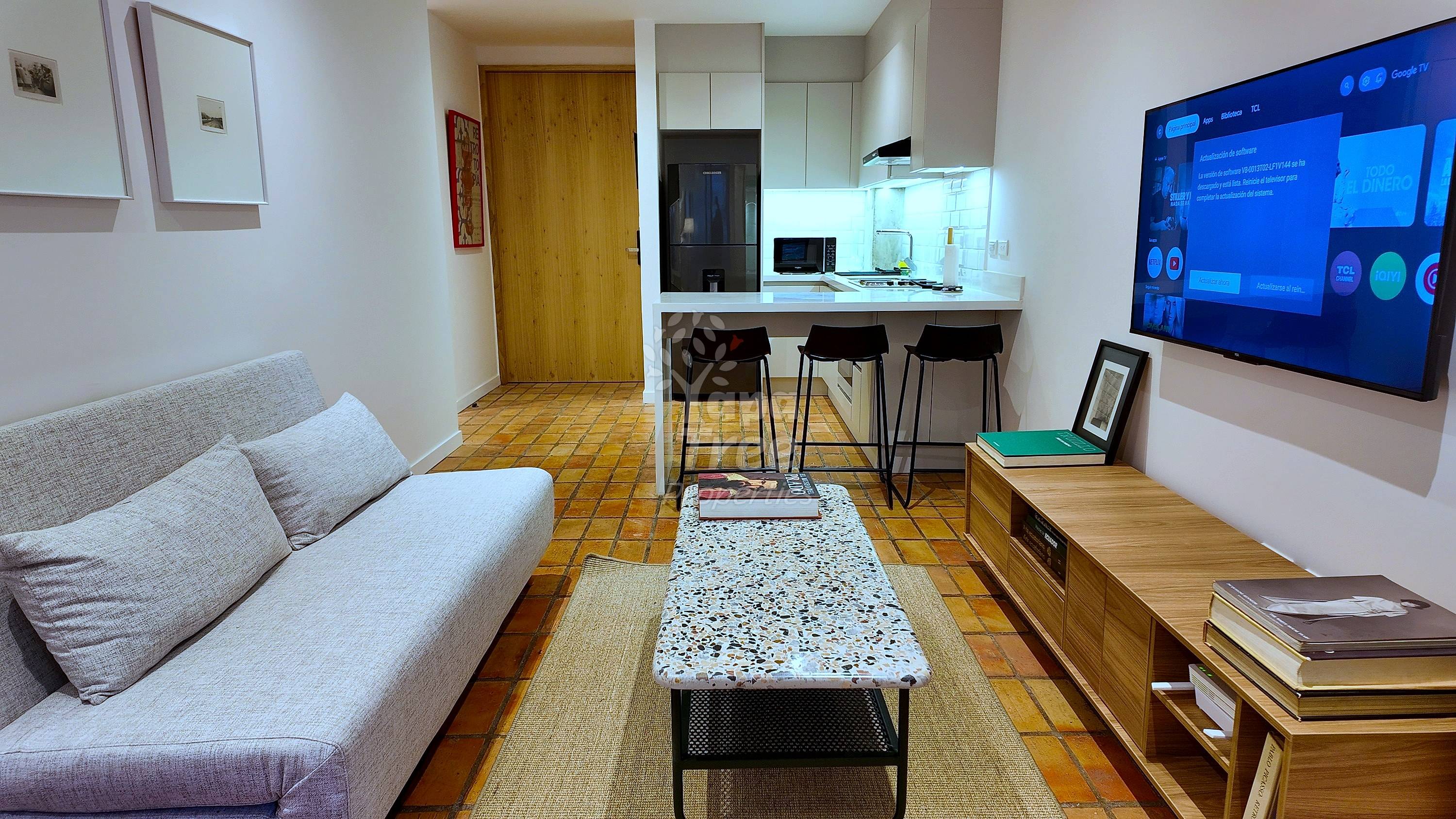 Área de estar en departamento de planta abierta con sofá gris, piso de baldosa terracota, paredes blancas, cocina con electrodomésticos negros y asientos de barra, televisor montado en pared, y muebles de madera.