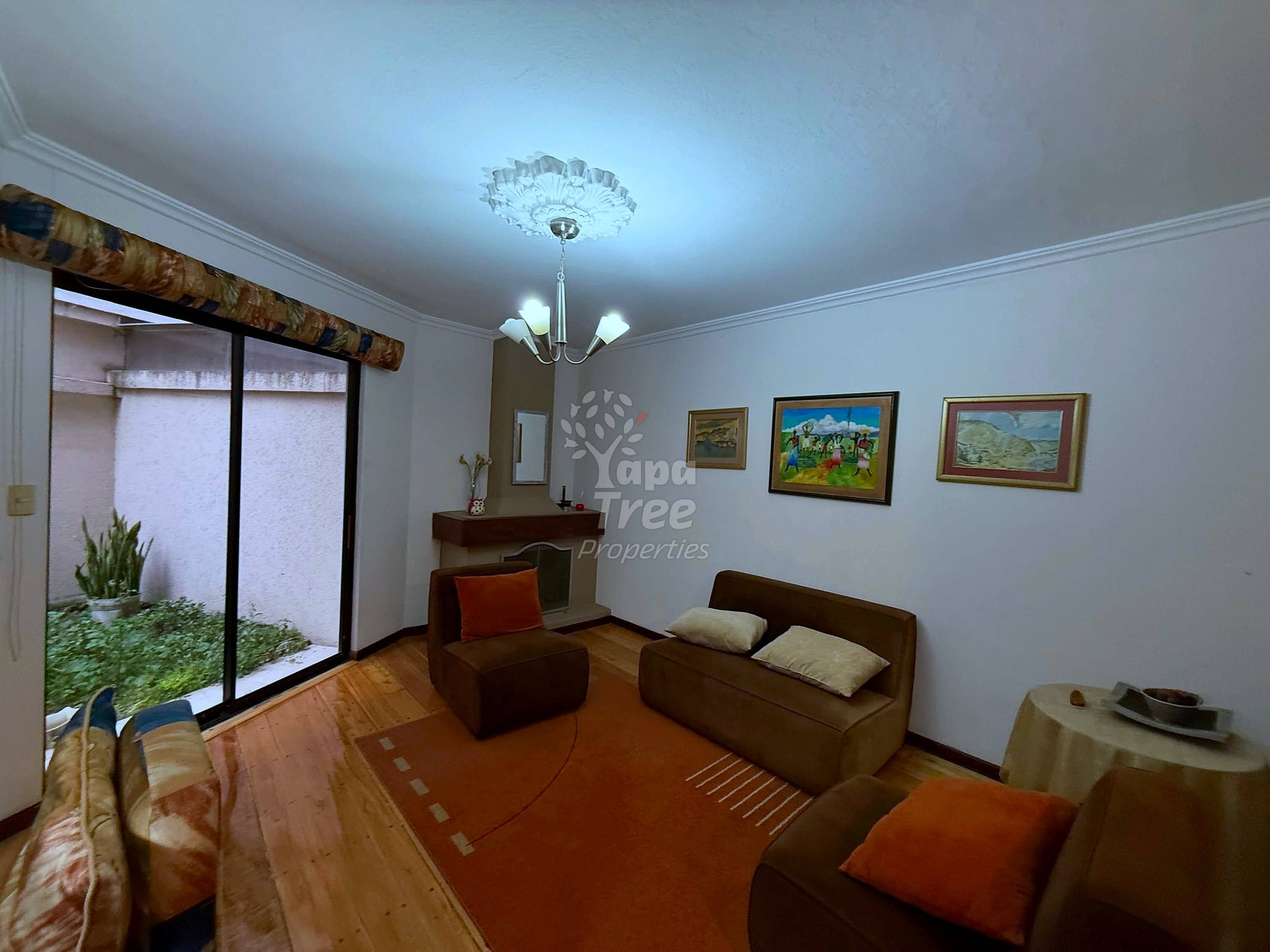 Living room with brown sofa, armchair, framed landscape paintings, chandelier, glass door opening to interior courtyard with plants.