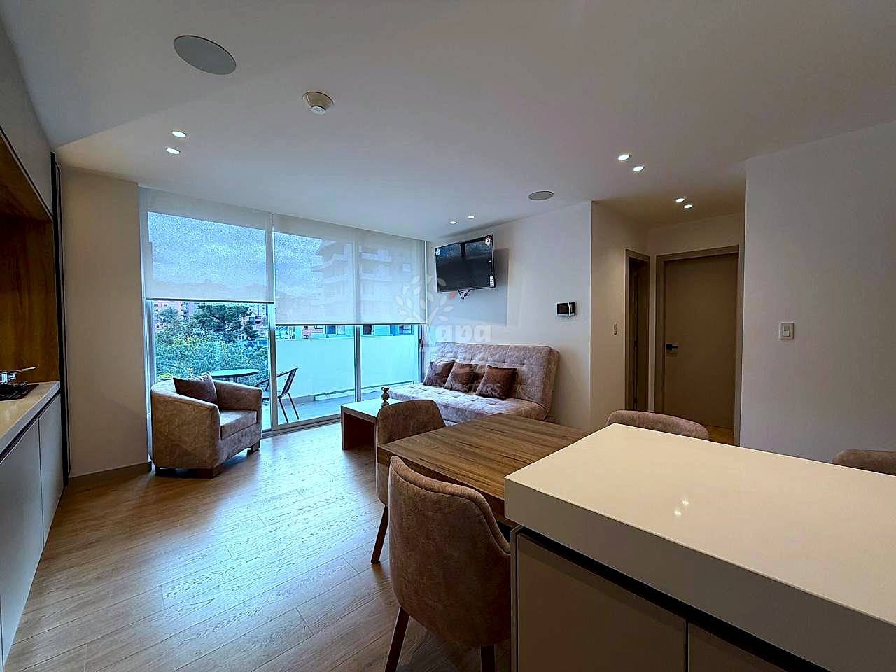 Open-plan departamento with white kitchen counter, brown upholstered sofa and chairs, wooden flooring, large windows with city views, and mounted television.