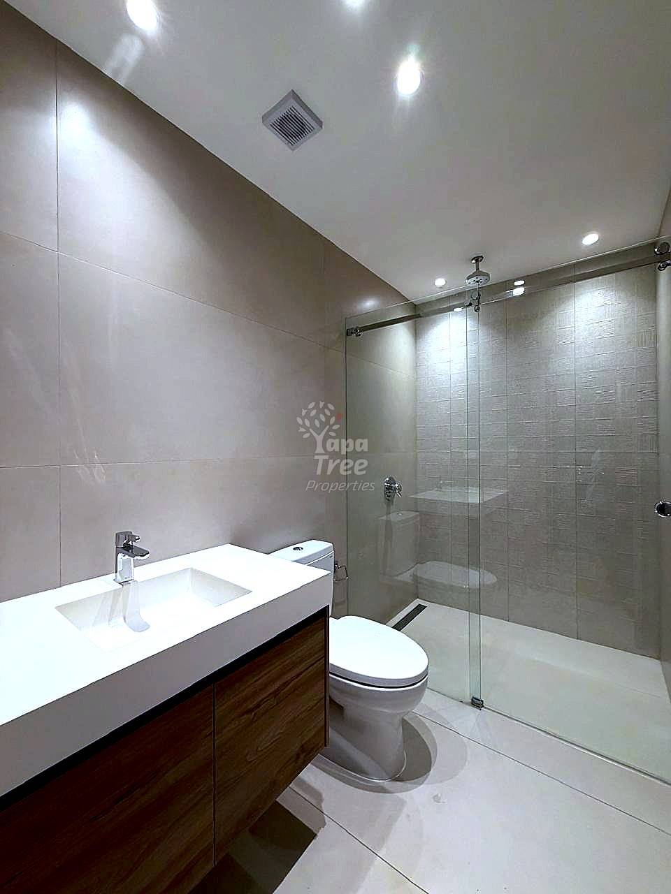 Modern bathroom with white tile walls, white countertop sink, dark wood vanity cabinet, white toilet, and glass-enclosed shower with overhead ventilation.