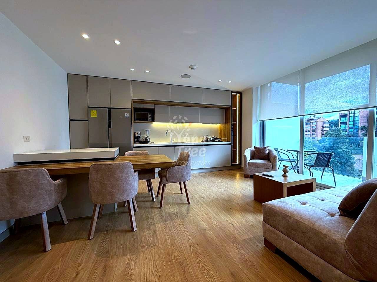 Open-plan departamento with modern gray kitchen, wooden dining table, beige upholstered seating, wood flooring, recessed lighting, and large windows with city views.