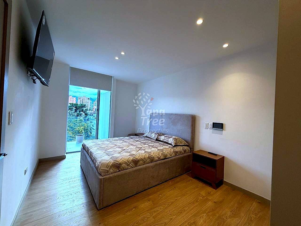 Bedroom with gray upholstered bed, wooden nightstand, wall-mounted TV, recessed ceiling lights, and glass door opening to city view balcony.