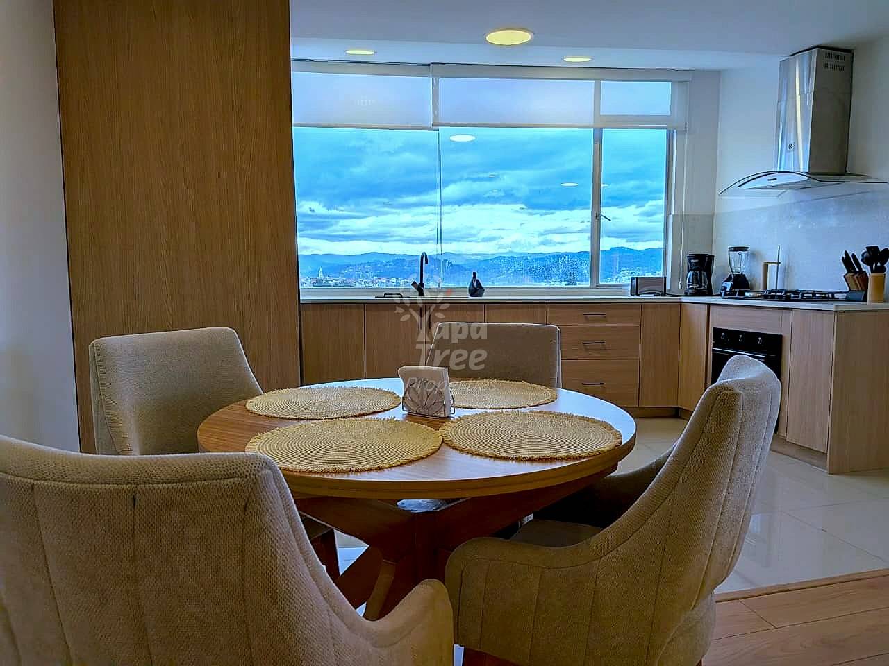 Open dining area with round wood table, four upholstered chairs, wood cabinetry, and large windows overlooking Cuenca valley with mountains.
