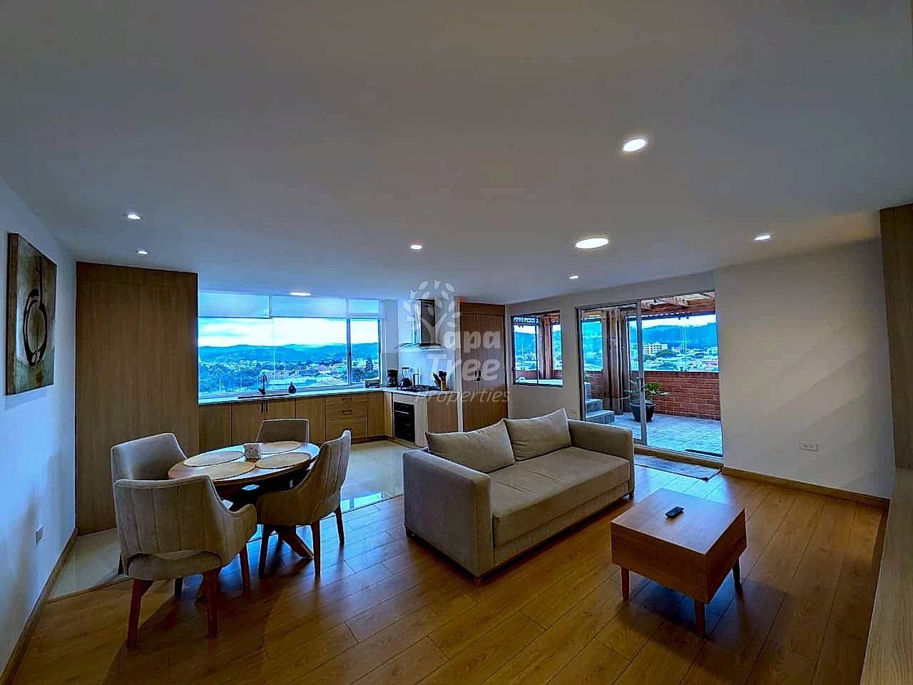 Open-concept living room and kitchen with wooden flooring, beige sofa, dining table, recessed ceiling lights, and mountain views through large windows.