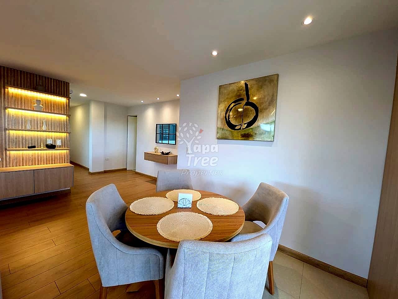 Dining area with round wooden table, four upholstered chairs, warm wood flooring, lit shelving unit, recessed ceiling lights, and abstract wall art.
