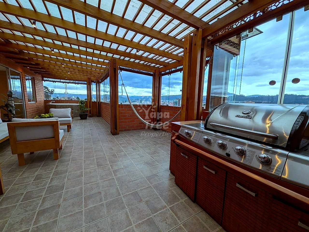 Covered rooftop terrace with wooden pergola, stainless steel grill, tile flooring, seating area, and panoramic city and mountain views through large windows.