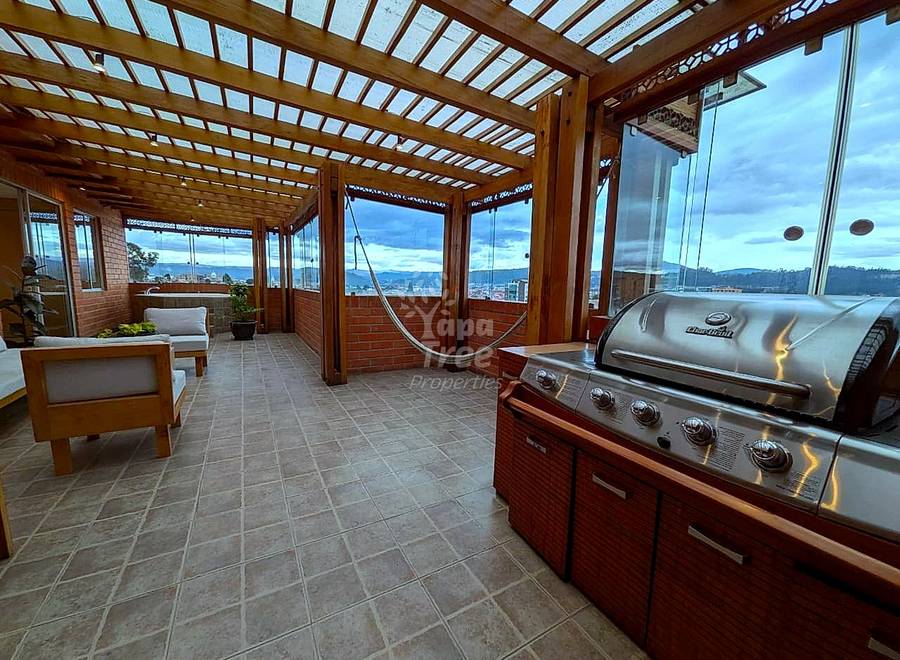 Covered rooftop terrace with wooden pergola, stainless steel grill, tile flooring, seating area, and panoramic city and mountain views through large windows.