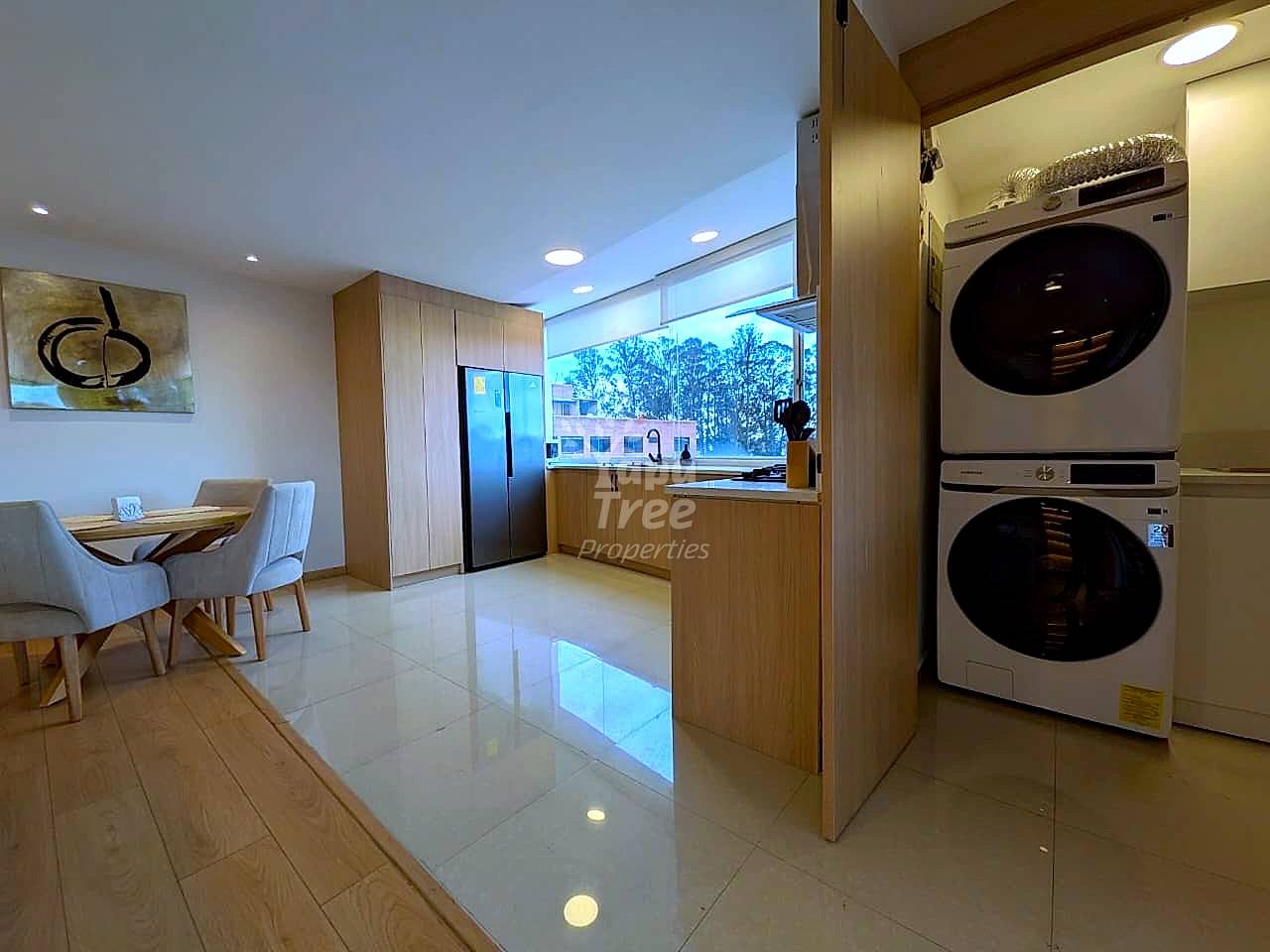 Modern kitchen and dining area with wooden cabinetry, black refrigerator, stacked dryer units, dining table with chairs, and polished tile flooring.