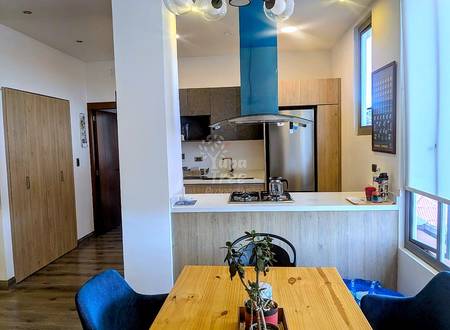 Modern kitchen with light wood cabinetry, stainless steel appliances, blue exhaust hood, and dining table with blue chairs in foreground.