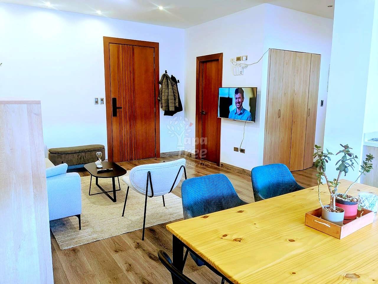 Open-plan living room with wooden dining table, blue upholstered chairs, light blue accent chair, wall-mounted TV, wooden doors, and recessed ceiling lights.