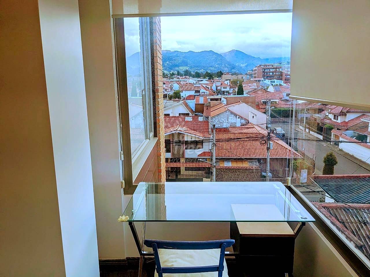 Home office with glass desk, blue chair, and floor-to-ceiling windows overlooking Cuenca's red-tile roofs and green mountains.