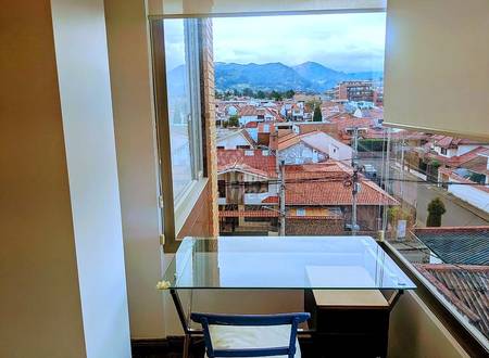 Home office with glass desk, blue chair, and floor-to-ceiling windows overlooking Cuenca's red-tile roofs and green mountains.