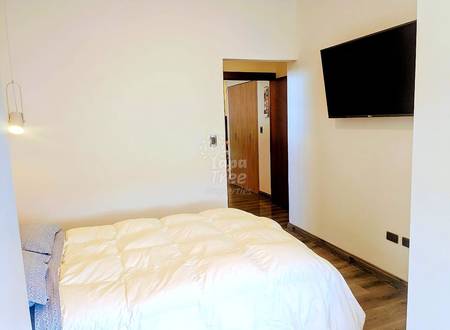Bedroom with queen bed, white bedding, wooden wardrobe, wall-mounted flat-screen TV, pendant light, and wood flooring.