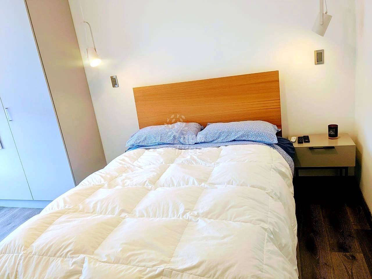 Bedroom with queen bed, wooden headboard, white duvet, blue pillows, wood nightstand, and white walls with recessed lighting.