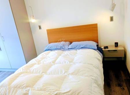 Bedroom with queen bed, wooden headboard, white duvet, blue pillows, wood nightstand, and white walls with recessed lighting.