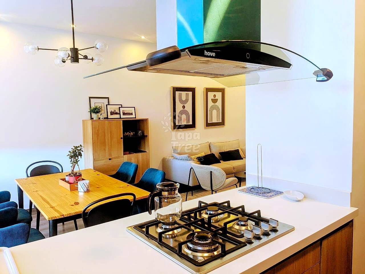Open-plan cocina and dining area with gas cooktop, white countertop, wooden table, and living room visible through opening with sofa and framed wall art.