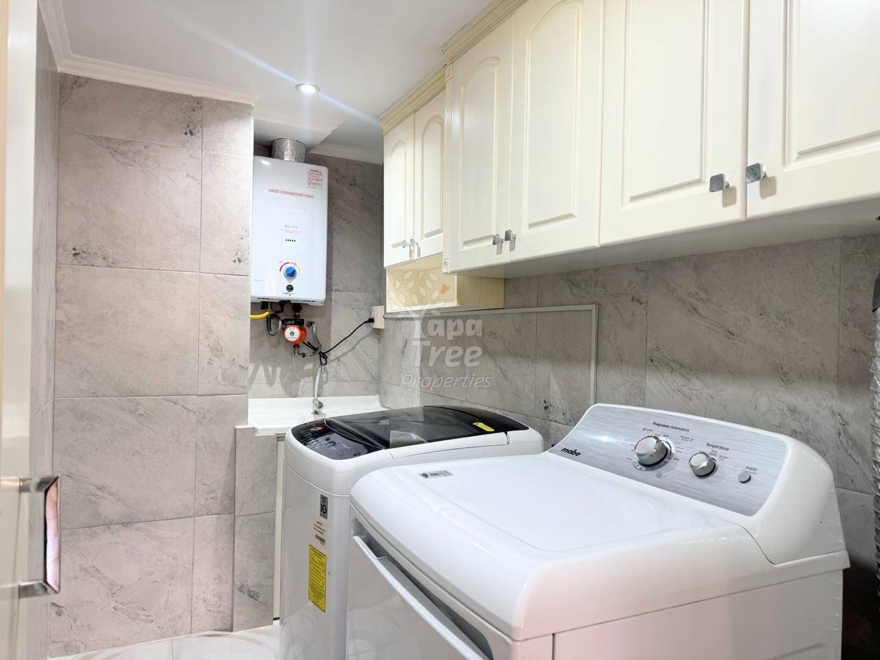Lavandería with white top-load washer, gray marble tile walls, cream cabinets, white tankless water heater, and gray quartz countertop.