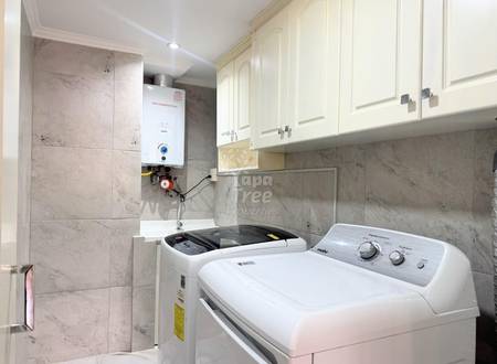 Lavandería con lavadora blanca de carga superior, paredes de azulejo mármol gris, gabinetes color crema, calentador de agua sin tanque blanco, y mesón de cuarzo gris.