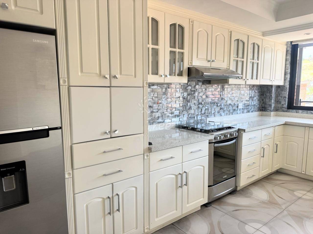 Modern cocina with white cabinetry, stainless steel appliances, gray tile backsplash, and light countertops with window access.