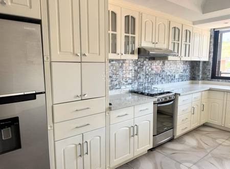 Cocina moderna con gabinetes blancos, electrodomésticos en acero inoxidable, salpicadero de azulejos grises y encimeras claras con acceso a ventana.