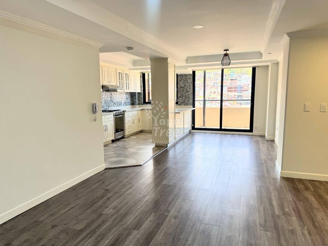 Sala abierta con piso de laminado tipo madera, paredes blancas, puertas de vidrio con marco negro hacia balcón, y cocina adyacente con gabinetes blancos y backsplash de azulejos.