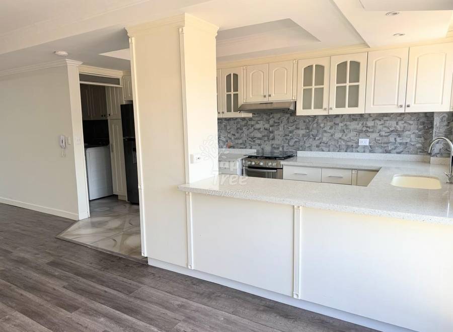 Cocina de concepto abierto con gabinetes blancos, isla con encimera de granito, backsplash de azulejos grises, estufa de acero inoxidable y piso tipo madera que se extiende al área de sala