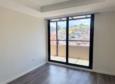 Habitación vacía con paredes color crema, piso de madera oscura, y puertas corredizas de vidrio con marco negro hacia balcón con vista a barrios de Cuenca.