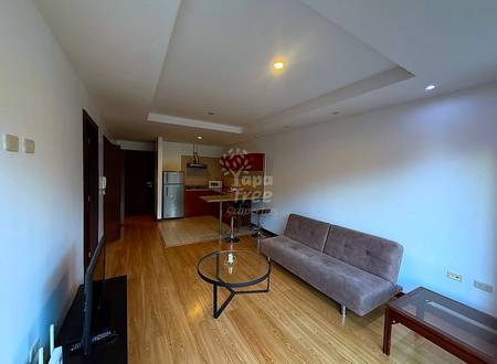 Open-concept departamento interior with wood flooring, gray sofa, round glass table, red kitchen with stainless steel refrigerator, and recessed ceiling lights.