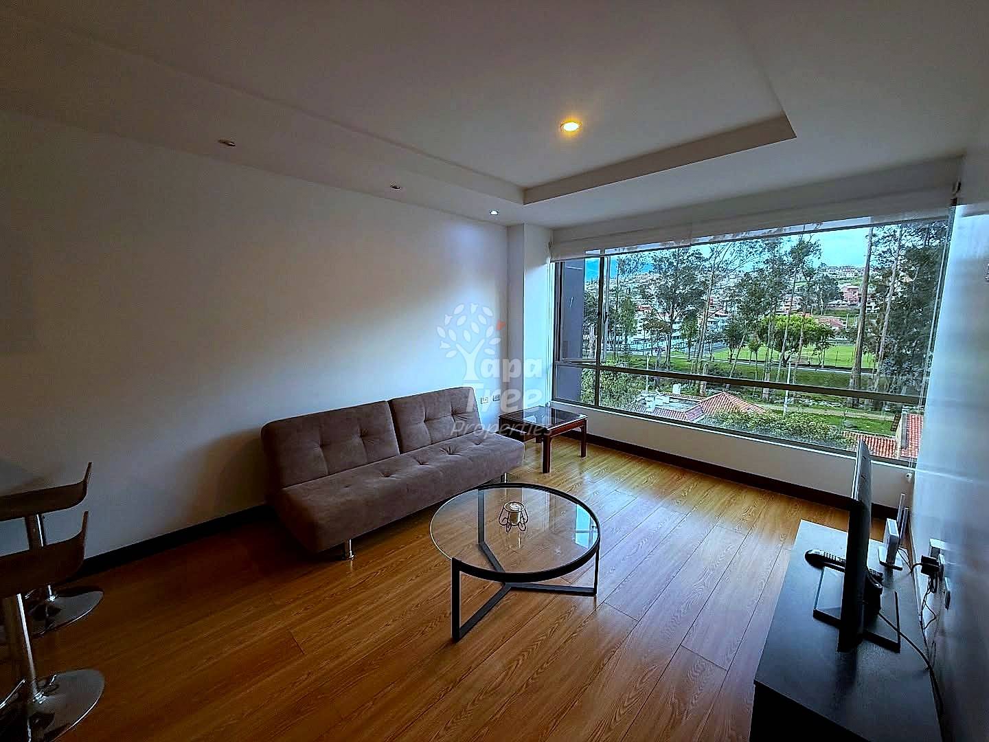 Living room with brown sofa, round glass table, wooden flooring, recessed lighting, and large windows overlooking park landscape.