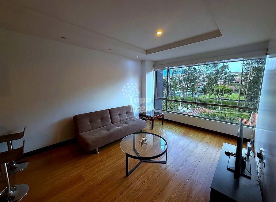 Living room with brown sofa, round glass table, wooden flooring, recessed lighting, and large windows overlooking park landscape.