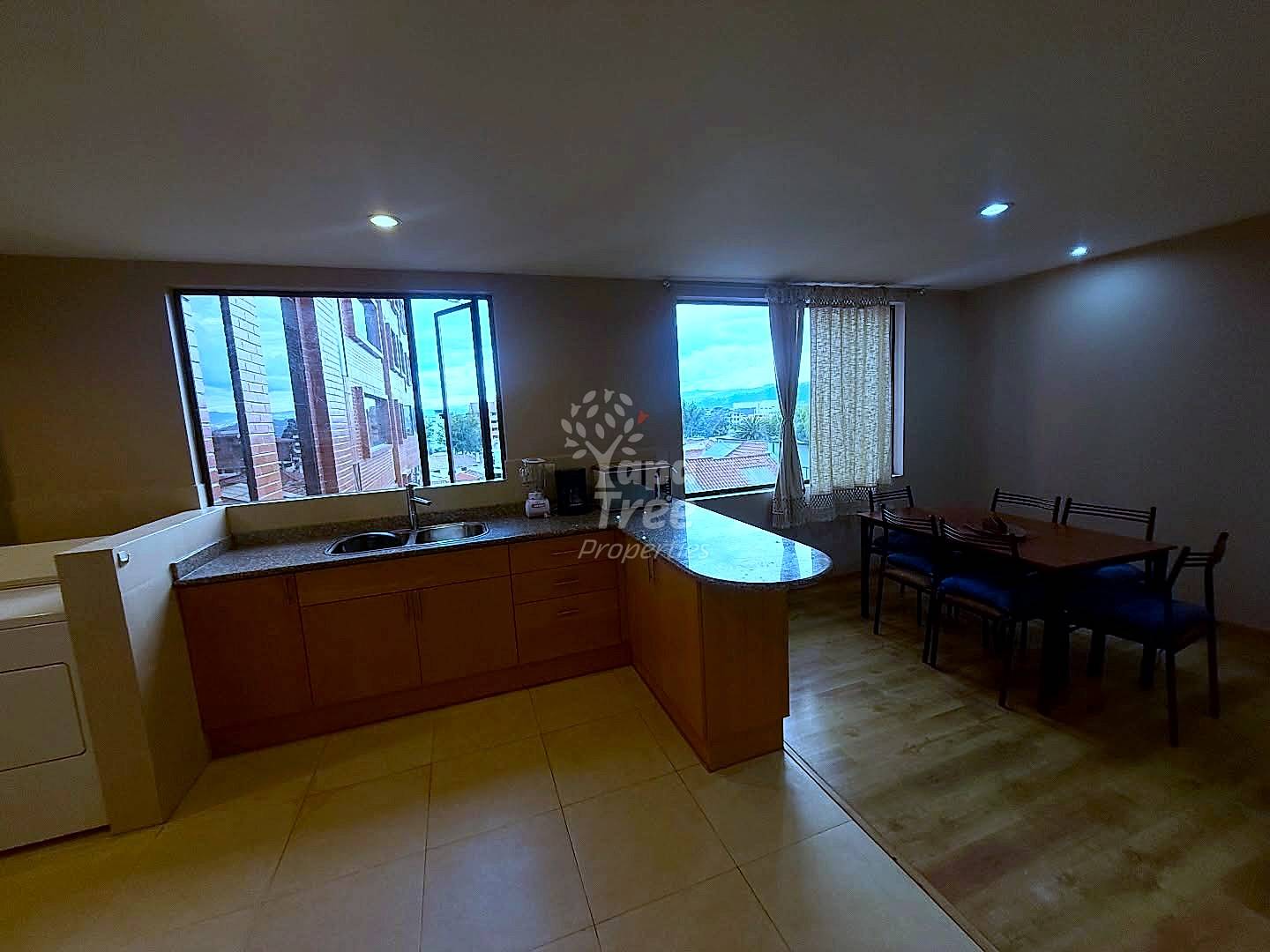 Open-plan kitchen and dining area with wood cabinetry, island counter, tile flooring, dining table with chairs, and two large windows with city views.