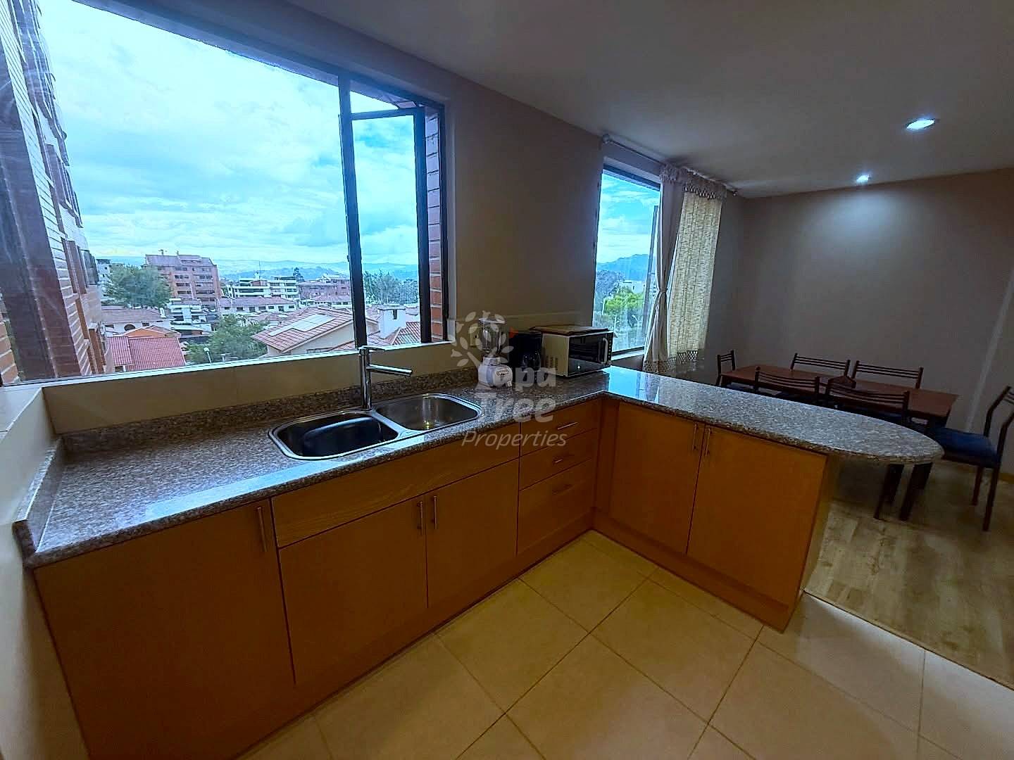 Kitchen with wooden cabinets, granite countertops, two sinks, and large windows overlooking Cuenca cityscape and mountains.