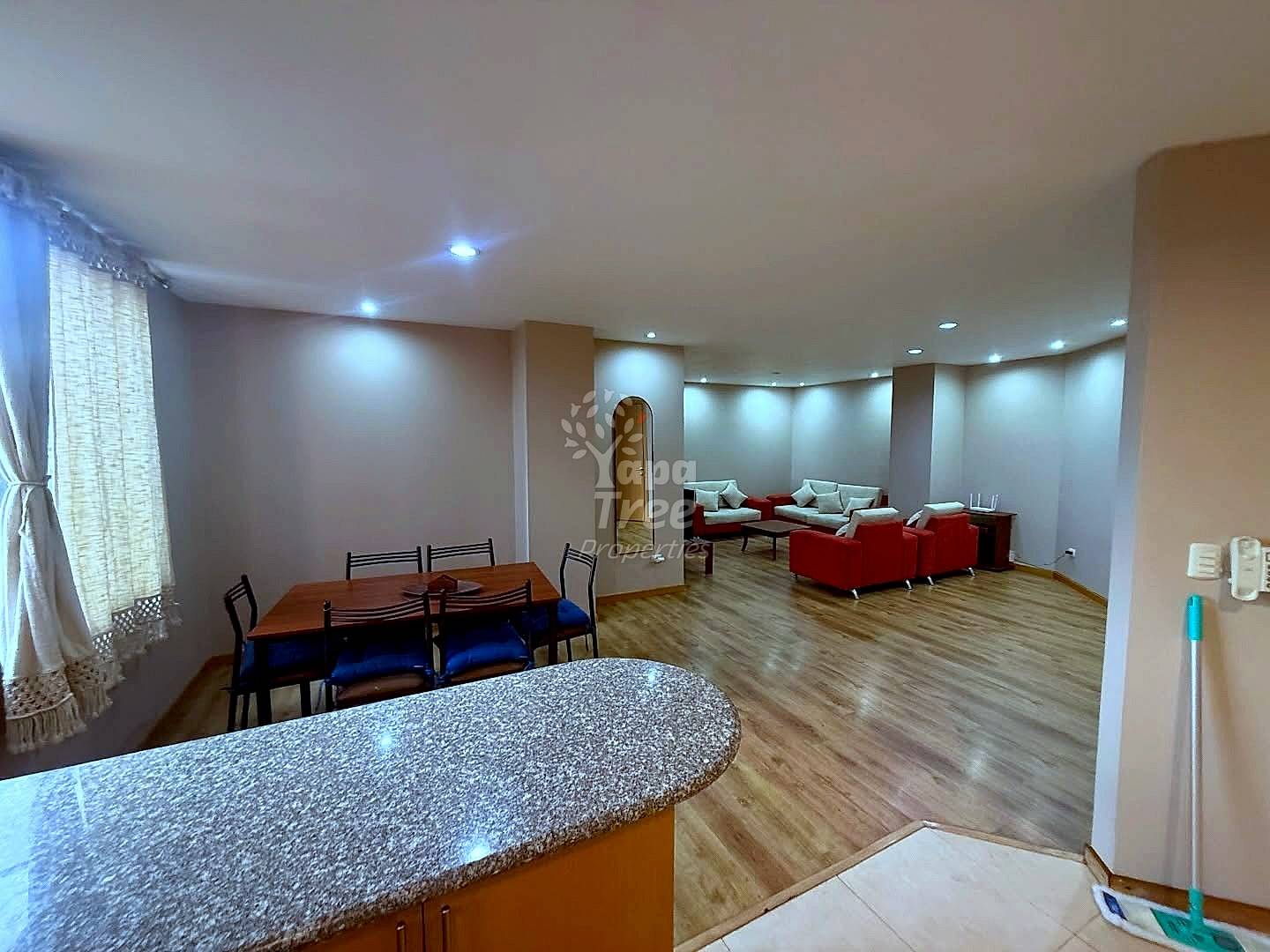 Open-concept departamento interior showing kitchen with granite countertop, dining area with wooden table, and living room with red and beige upholstered seating, recessed lighting, wood laminate flooring throughout.
