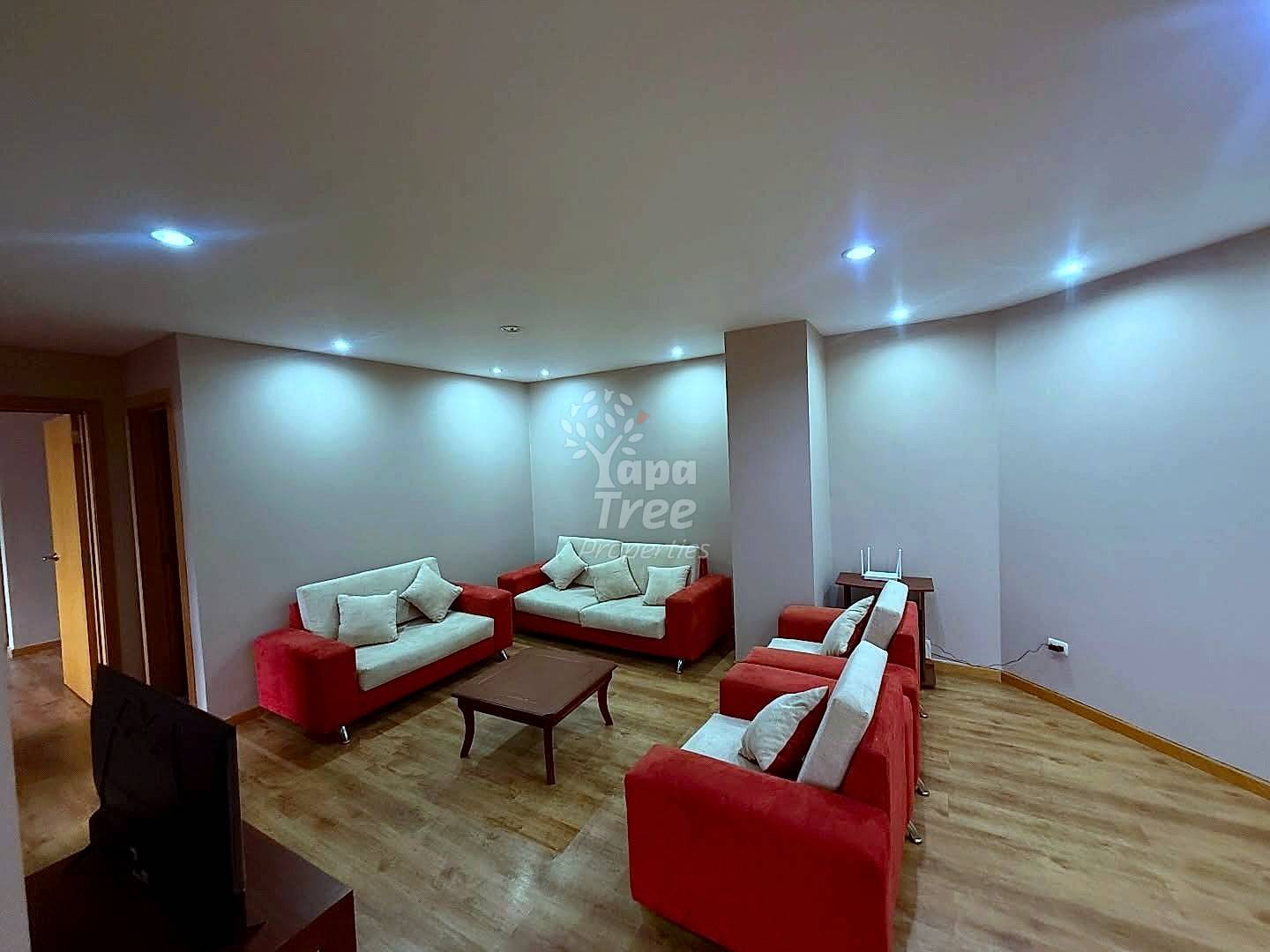 Living room with three red and cream sofas, wooden coffee table, recessed ceiling lights, beige walls, and wooden flooring throughout.