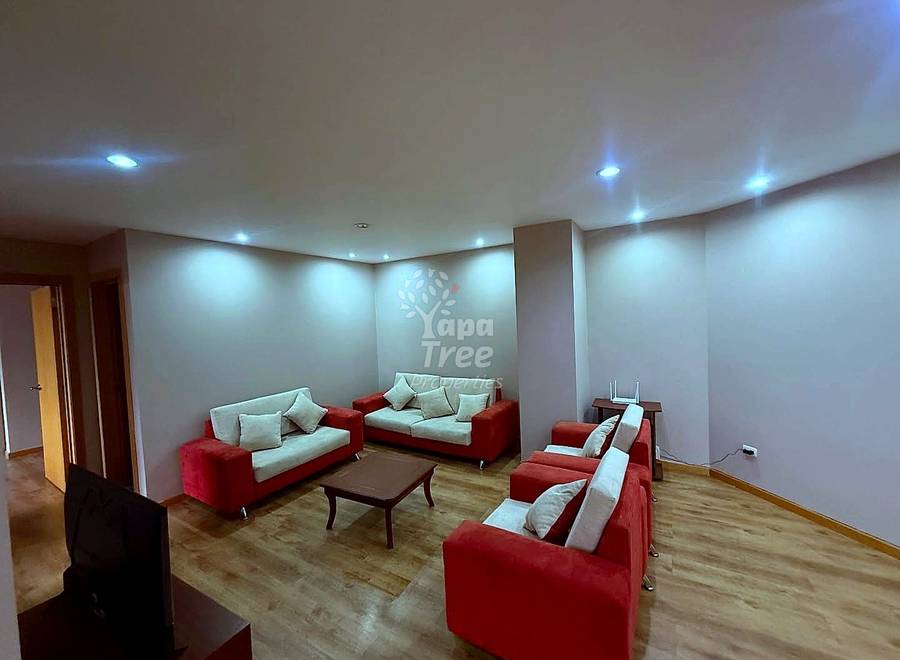Living room with three red and cream sofas, wooden coffee table, recessed ceiling lights, beige walls, and wooden flooring throughout.