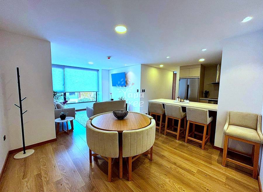 Open-plan departamento interior with round wooden dining table, upholstered chairs, hardwood flooring, recessed ceiling lights, kitchen counter with bar seating, and window with blinds.