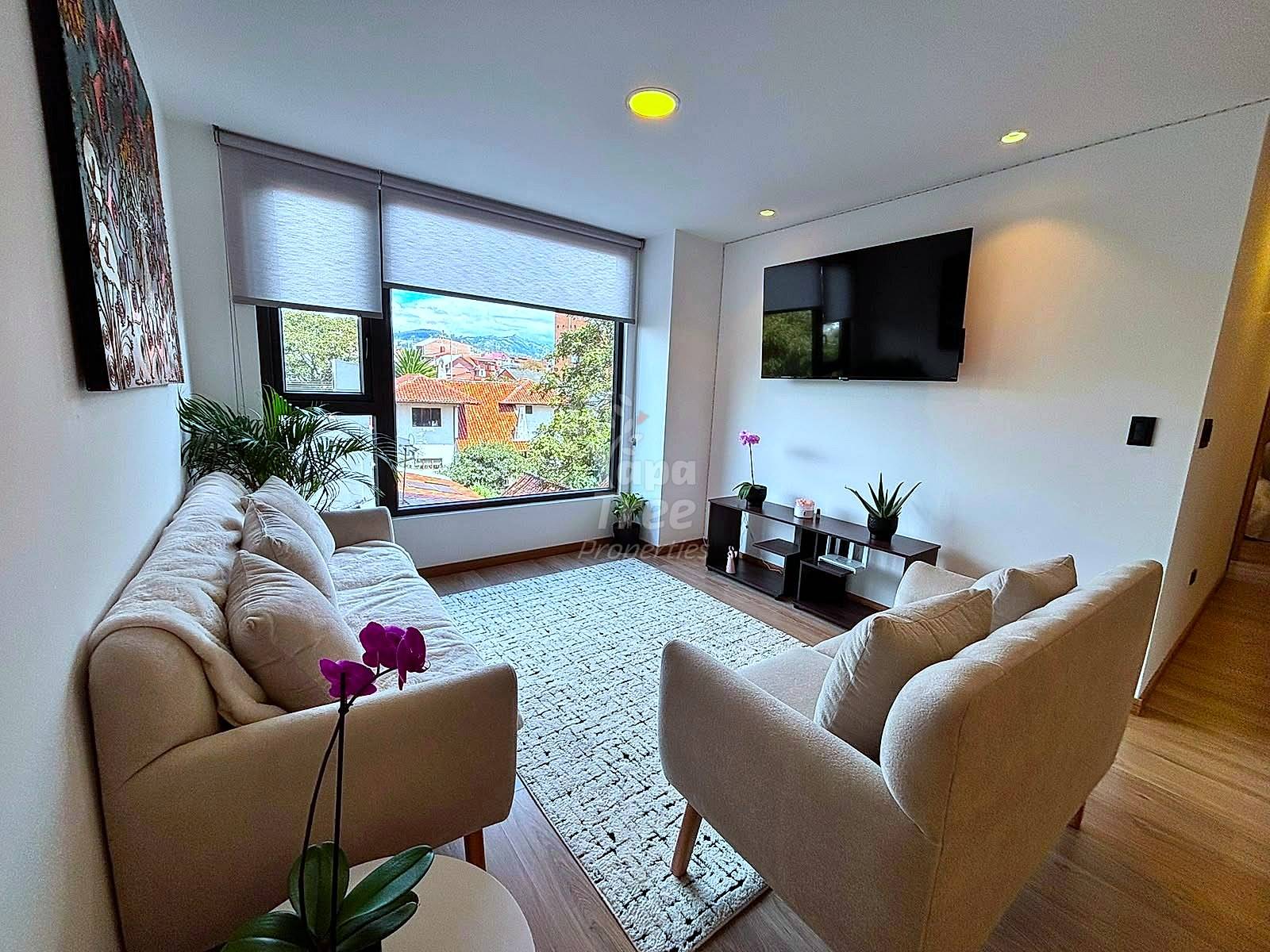 Living room with beige sofa and armchair, wall-mounted TV, large windows with city views, recessed ceiling lights, wooden flooring, and potted plants.