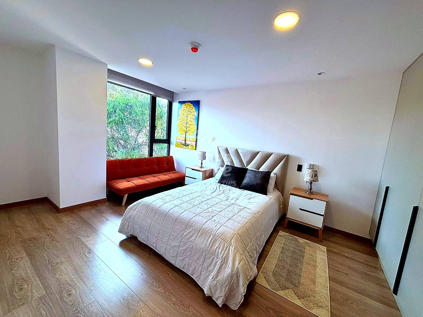 Bedroom with queen bed, beige upholstered headboard, wood nightstand, orange sofa by window, recessed ceiling lights, wood flooring, and framed artwork.