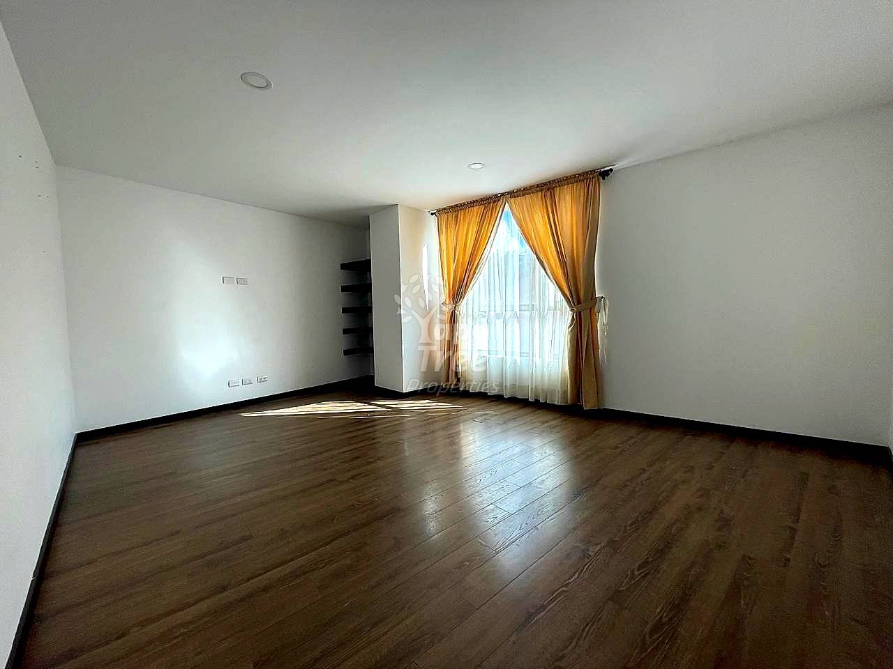 Empty living room with white walls, recessed ceiling lights, dark wood flooring, and large window with golden curtains providing natural light.