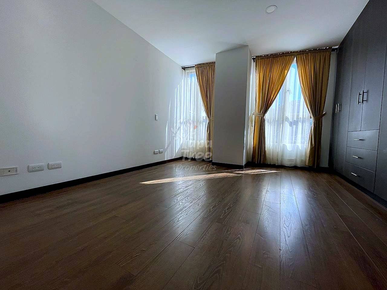 Empty bedroom with dark wood flooring, white walls, two windows with tan curtains, gray built-in closet, and natural light.