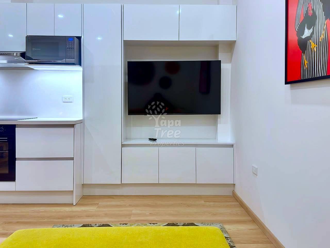 Modern open-concept living area with white cabinetry, wall-mounted flat-screen TV, microwave, wooden flooring, and yellow accent furniture.
