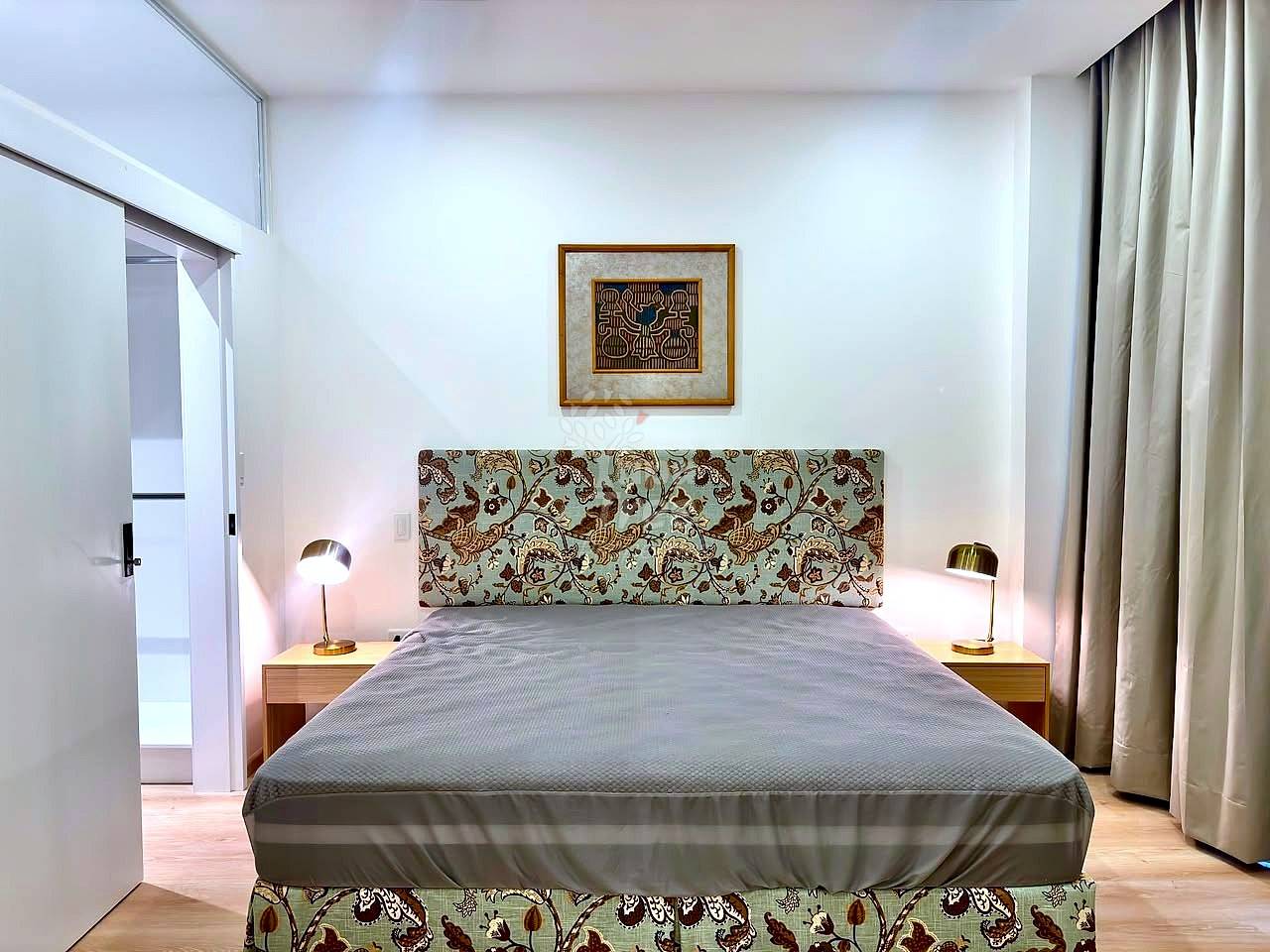 Master bedroom with queen bed, patterned headboard, two wooden nightstands with brass lamps, framed artwork, white walls, and cream curtains.